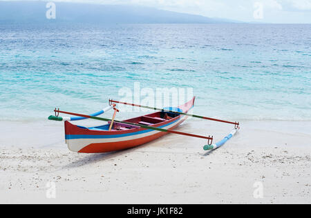 Rot weiß blau gefärbte kleine leere Kanu Boot gestrandet auf einem tropischen Sandstrand mit blauen Ozeanwasser an einem sonnigen Tag am Nachmittag. Stockfoto