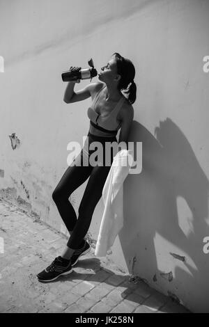 Müde junge Sportlerin stützte sich auf Wand im Stadion und Trinkwasser aus Sport-Flasche, schwarz / weiß-Bild Stockfoto