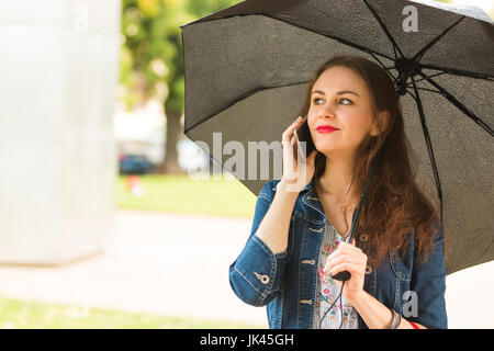 Junge Frau hält Regenschirm und reden über Handy im Freien an regnerischen Tag Stockfoto