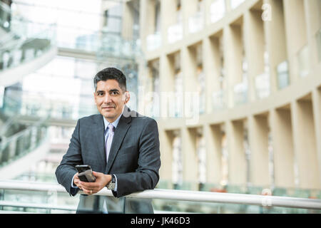 Gemischte Rennen Geschäftsmann stützte sich auf Geländer in Lobby hält Handy Stockfoto