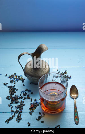 vertikale Schuss von Kräuter-Tee-Glas mit Sieb, Nahost dekorative Kanne, lebendige Teelöffel und Tee Blätter auf blauem Hintergrund. erhöhte Ansicht. viele Stockfoto
