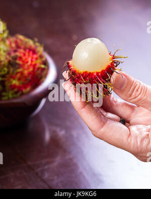 Nahaufnahme einer Frau Hand hält eine Rambutan, die offen in der Nähe von einer Schüssel mehr diese köstliche Frucht geschnitten worden Stockfoto