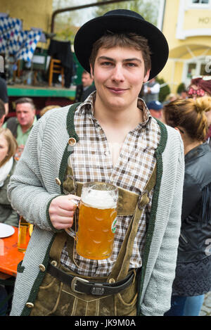 Porträt eines lächelnden jungen bayerischen Mannes in bayrischer Tracht mit Lederhosen, Filz schwarz, Hut, graue Strickjacke, braun checked Shirt und Bier Becher Stockfoto