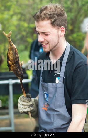 Junger Mann runzelte die Stirn und hält einen Fisch am Stiel wie Forelle, Saibling oder Felchen gegrillt über glühende Kohle in ein traditionelles Barbecue Essen in Bayern Stockfoto