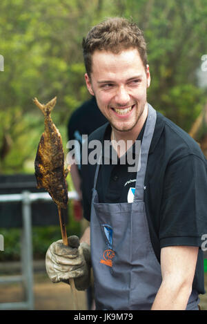 Junger Mann, lachend und hält einen Fisch am Stiel wie Forelle, Saibling oder Felchen gegrillt über glühende Kohle in ein traditionelles Barbecue Essen in Bayern Stockfoto