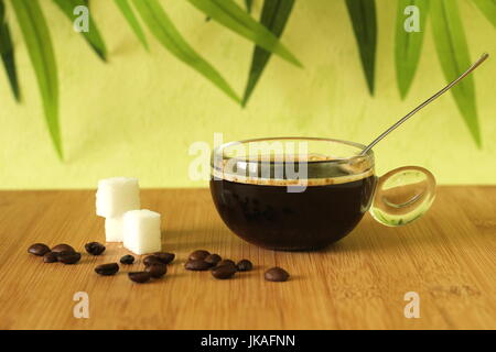 Eine Tasse schwarzen Kaffee auf braunem Bambus Holzboden mit Stücken von Zucker gestapelt neben ihm und Kaffeebohnen auf grünem Laub Hintergrund Stockfoto