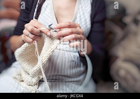 Eine junge Frau strickt ein gestreiftes Kleid mit weißer Wolle stricken wolle einen warmen Winter Pullover strickt und sitzt zu Hause auf der Couch Stockfoto