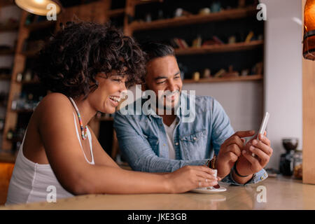 Junge glückliche Paar am Tisch im Café sitzen und mit Blick auf Handy und lächelnd. Afrikanerin und kaukasischen Mann in Coffee-Shop mit Smartphone. Stockfoto