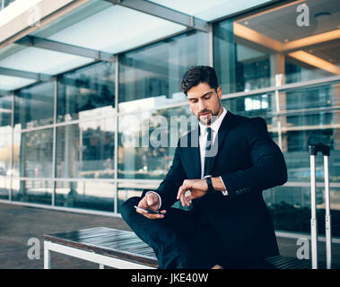 Junger Geschäftsmann warten und überprüfen mit Handy. Geschäftlich unterwegs, auf Bank sitzend und mit Blick auf seine Uhr am Flughafen-terminal. Stockfoto