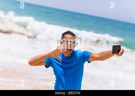 Mann mit Sonnenbrille sitzt mit einem Smartphone in den Händen und machen Selfie am Telefon in der Nähe des Meeres Stockfoto