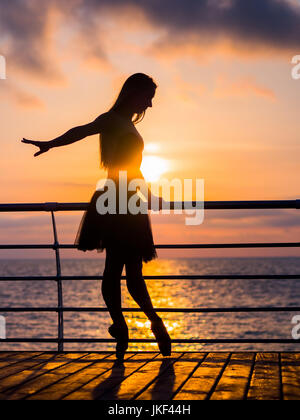 Silhouette der tanzende Ballerina in schwarz Ballett Tutu und Pointe auf Damm über dem Ozean oder Meer bei Sonnenaufgang oder Sonnenuntergang. Junge attraktive blonde Frau w Stockfoto