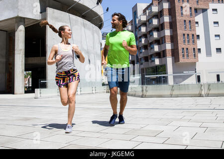 Gut aussehender Mann und schöne Frau, die zusammen auf der Straße zwischen Wohnhäusern am sonnigen Tag joggen Stockfoto