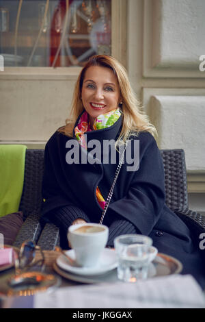 Frau sitzt in einem Restaurant, Kaffee trinken und wegsehen links von ihr mit einem Lächeln, als sie etwas mit dem unteren Gesicht verdeckt Cup sieht Stockfoto