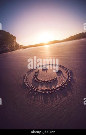 Lächelndes Gesicht auf einem sandigen Strand mit Sonnenuntergang im Hintergrund gezeichnet. Stockfoto