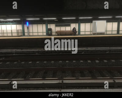 Mann in Hoodie sitzt auf Holzbank Blick auf sein Handy warten auf die u-Bahn in der Nacht. Spät in die Nacht warten auf die New Yorker u-Bahn zu trainieren. Stockfoto