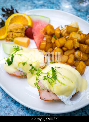 Klassische Eggs Benedict (weiche Spiegeleiern, kanadische zurück Speck und Sauce Hollandaise auf eine englische Muffins), gebratene Kartoffeln und Obst zum Frühstück. Stockfoto