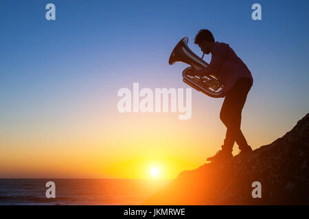 Tuba - Instrument. Silhouette eines jungen Mannes, der Trompete auf felsigen Küste während des Sonnenuntergangs. Stockfoto