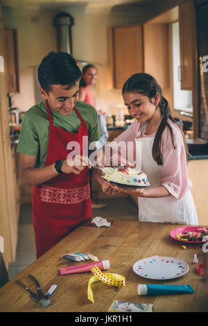 Bruder und Schwester dekorieren einen Kuchen zusammen in der Küche ihres Hauses. Ihre Mutter ist unscharf im Hintergrund erkennbar. Stockfoto
