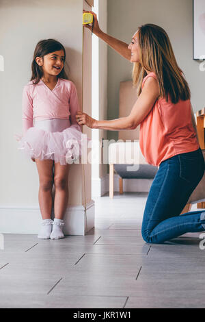 Kleines Mädchen in einem Ballett Kostüm mit ihrer Höhe Maß von ihrer Mutter. Sie steht an der Wand, während ihre Mutter das Maßband verwendet. Stockfoto