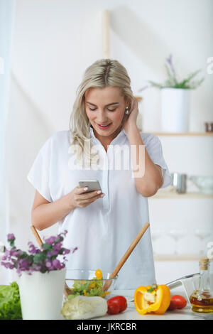 Lächelnde junge Frau mit Handy während der Vorbereitung Salat am Küchentisch Stockfoto