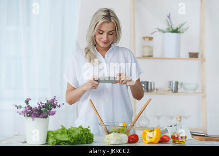 Lächelnde junge Frau fotografieren frischen Salat durch Smartphone in Küche Stockfoto