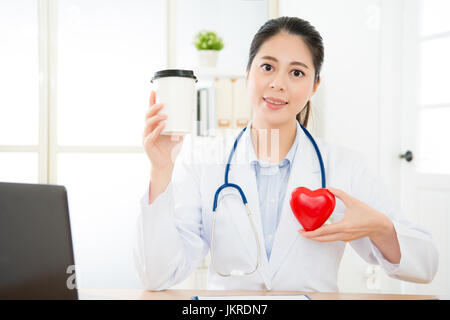 lächelnden jungen weiblichen Kardiologie Arzt Gesicht zur Kamera auf Schreibtisch und halten Kaffee Tasse zeigt Koffein verursacht Herzklopfen Problem Konzept. Stockfoto