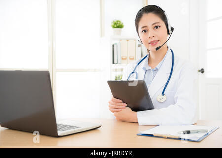 Ärztin, halten ihre digital-Tablette in hellen modernen Krankenhaus sitzen und sprechen mit ihren Patienten online mit Computer geben professionelle dient Stockfoto