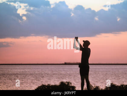 Ein junger Mann nimmt ein Selbstporträt ist gegen einen Sonnenuntergang am See Silhouette. Stockfoto