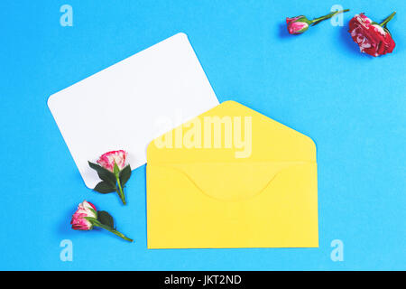 Leere Grußkarte mit gelben Umschlag und rosa Rosen-Blumen auf blauem Hintergrund. Ansicht von oben Stockfoto
