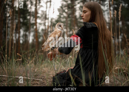 Mädchen mit langen Haaren in roten und schwarzen Kleid sitzt auf dem Rasen mit Eule auf der Hand im Wald. Eule ist durch Kette an den Arm gebunden. Stockfoto