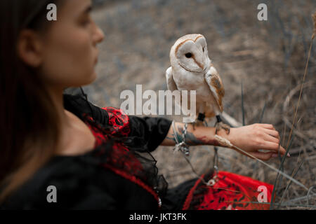 Mädchen im roten und schwarzen Kleid sitzt auf dem Rasen mit Eule auf der Hand im Wald. Eule ist durch Kette an den Arm gebunden. Close-up. Stockfoto