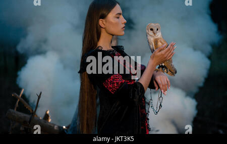 Mädchen mit langen Haaren in roten und schwarzen Kleid steht mit Eule auf der Hand im Wald vor Rauch Hintergrund. Eule ist durch Kette an den Arm gebunden. Stockfoto