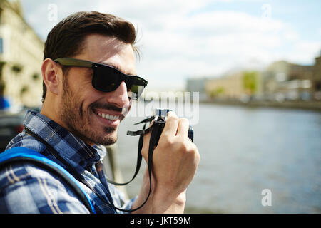 Porträt von gut aussehenden jungen Mann tragen Sonnenbrillen lächelnd Blick in die Kamera beim Fotografieren am Ufer des Flusses touristische Reise in den Urlaub zu genießen Stockfoto