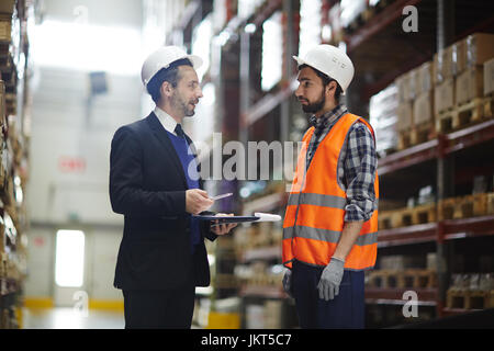Porträt der Lagerleiter Business Anzug im Gespräch mit Lader im Gang zwischen hohen Regalen mit verpackter Ware Stockfoto