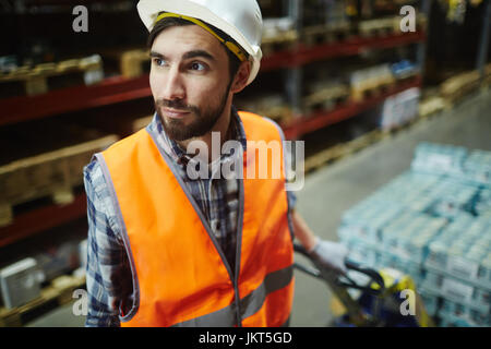 Porträt des bärtigen Loaders arbeiten im Lager, ziehen bewegten Karren mit Handelswaren im Gang zwischen hohen Regalen Stockfoto