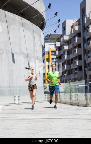 Gut aussehender Mann und schöne Frau, die zusammen auf der Straße zwischen Wohnhäusern am sonnigen Tag joggen Stockfoto