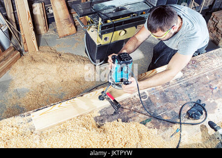 Ein starke männliche Schweißer in Arbeit Kleidung mit Begeisterung arbeiten und sägt ein Holzbrett mit einer Kreissäge auf einem Holztisch in einem industriellen Workshop ein Stockfoto