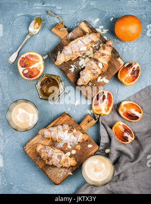 Traditionellen italienischen Stil zu Hause Frühstück. Latte in Gläsern, blutige Mandel Croissants auf urigen Holzbrettern und rot sizilianischen Orangen über strukturierte Betontisch Stockfoto