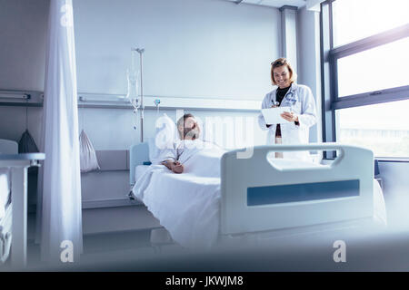 Ärztin im Gespräch mit männlichen Patienten im Krankenhausbett. Lächelnde Arzt mit Zwischenablage kranken Mann im Krankenhaus besucht. Stockfoto