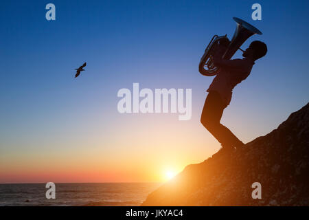 Silhouette eines jungen Mannes, der Trompete auf felsigen Küste bei traumhaften Sonnenuntergang. Tuba-Instrument. Stockfoto