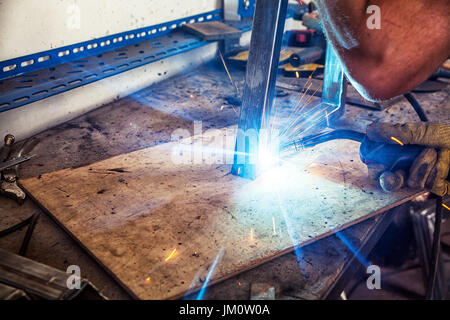 Eine Nahaufnahme des Schweißens ein Metal Ecke mit einem Schweißgerät auf einem Holztisch in der Fabrik, blaue Funken fliegen auseinander Stockfoto