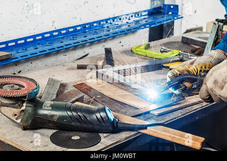 Close-up einen erwachsenen Mann im schützenden Bau Handschuhe und Kochen ein Metall Winkel mit einem Schweißgerät auf einem Holztisch in der Fabrik, um eine di Stockfoto