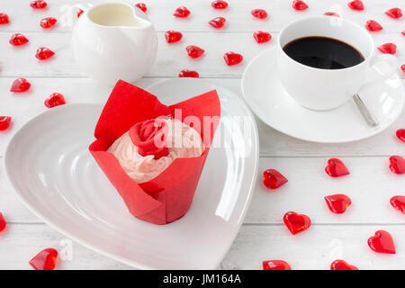 Valentines Cupcake dekoriert mit einer roten Rose auf einem weißen Teller mit einer Tasse Kaffee und Milch Krug im Hintergrund. Weißen hölzernen Hintergrund geschmückt w Stockfoto