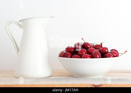 Schale mit Kirschen als nächstes an eine rustikale Metall Glas auf einem weißen Holzregal. Leere Kopie Platz für Text Editor. Stockfoto