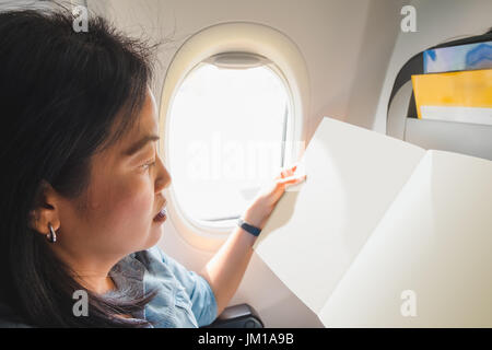 Asiatische Frau sitzt am Fenster Platz im Flugzeug und bei leeren offenen Broschüre lesen, wenn Sie fliegen, verspotten für Ihr Design hinzufügen. Stockfoto