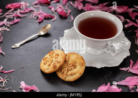 Schwarzer Kaffee oder Tee in weiße Tasse mit zwei Erdnusskekse und Pfingstrose Blütenblätter verstreut Stockfoto