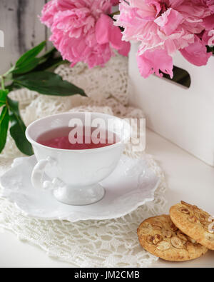 Rote Beeren oder Früchte Tee in weißem Porzellan Teetasse auf einem weißen Tisch mit Pfingstrose Bouquet und Cookies. Mütter Tag Konzept Stockfoto