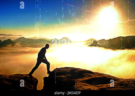Film-Korn-Effekt.  Wanderer in schwarz springt zwischen den felsigen Gipfeln. Wunderbare Götterdämmerung im Felsengebirge, schwere orange Nebel im tiefen Tal. Miracl Stockfoto