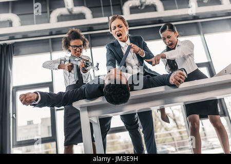 Drei junge wütend Geschäftsfrauen mit Gewehr Geschäftsmann auf Tisch, Business Team Meeting Konzept zu bestrafen Stockfoto