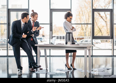 Zwei lachende Geschäftsleute zeigen mit Fingern auf gestresste Geschäftsfrau im Büro Stockfoto